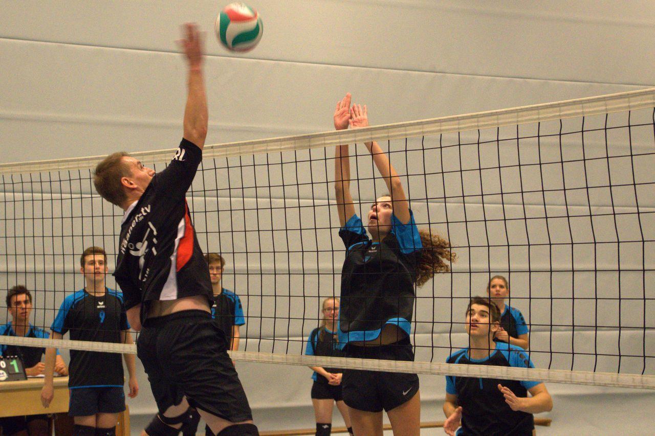 Volleyballspiel: Ein Spieler springt zum Block, während ein anderer in der Hintergrund zusieht. Ball über Netz.