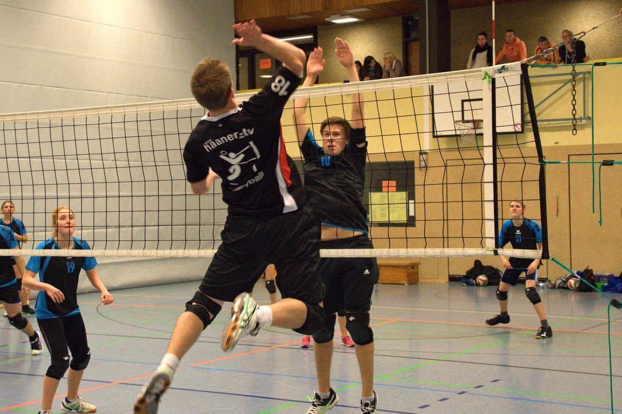 Zwei Volleyballspieler im Sprung am Netz, während Zuschauer im Hintergrund beobachten. Ein Spieler trägt ein schwarzes Trikot.