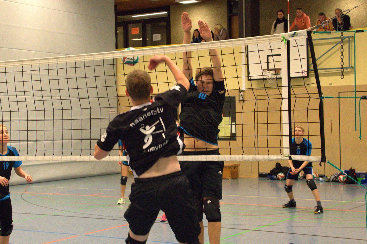 Spielszene beim Volleyball: Ein Spieler springt zum Aufschlag, während ein anderer blockt. Zuschauer sind im Hintergrund zu sehen.