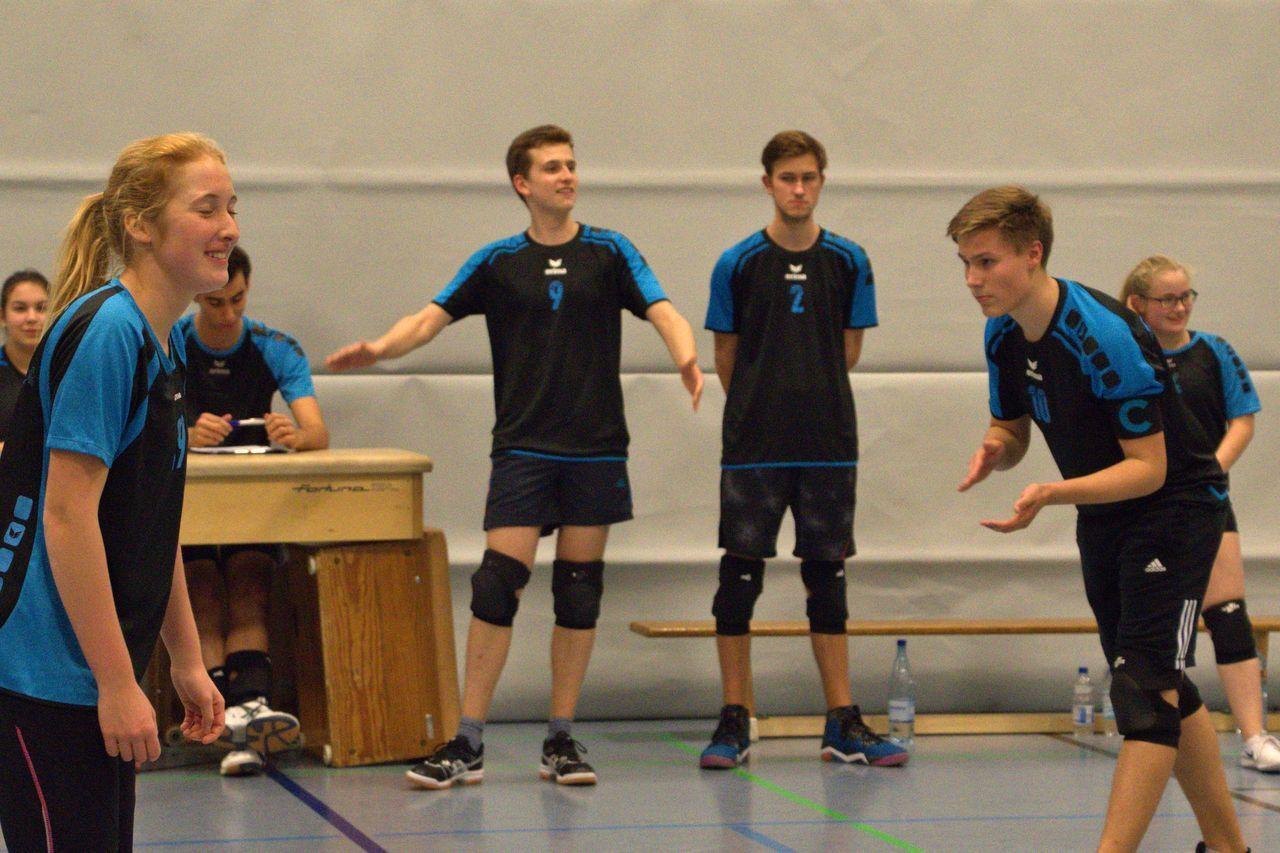 Eine Gruppe von Volleyballspielern in Schwarz-Blau interagiert auf dem Spielfeld, während eine Person an der Bank sitzt.