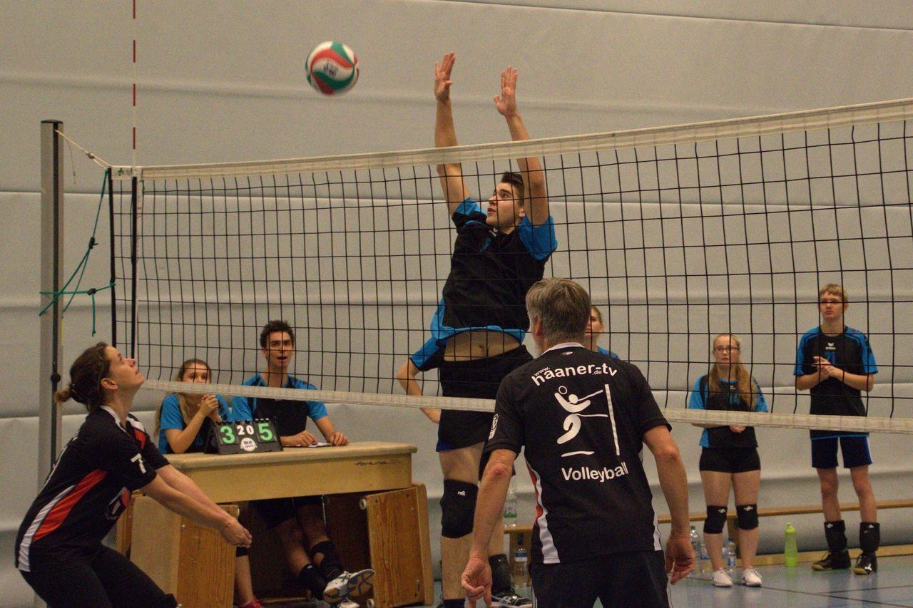 Spielszene im Volleyball: Eine Spielerin hebt den Ball zur Annahme, während ein Spieler am Netz springt. Zuschauer beobachten gespannt.