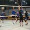 Volleyballspiel in der Halle: Spieler springen, um einen Ball über das Netz zu blocken, während andere zuschauen.