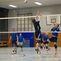 Volleyballspiel in einer Halle, ein Spieler springt, um den Ball über das Netz zu schlagen, während andere Zuschauer beobachten.