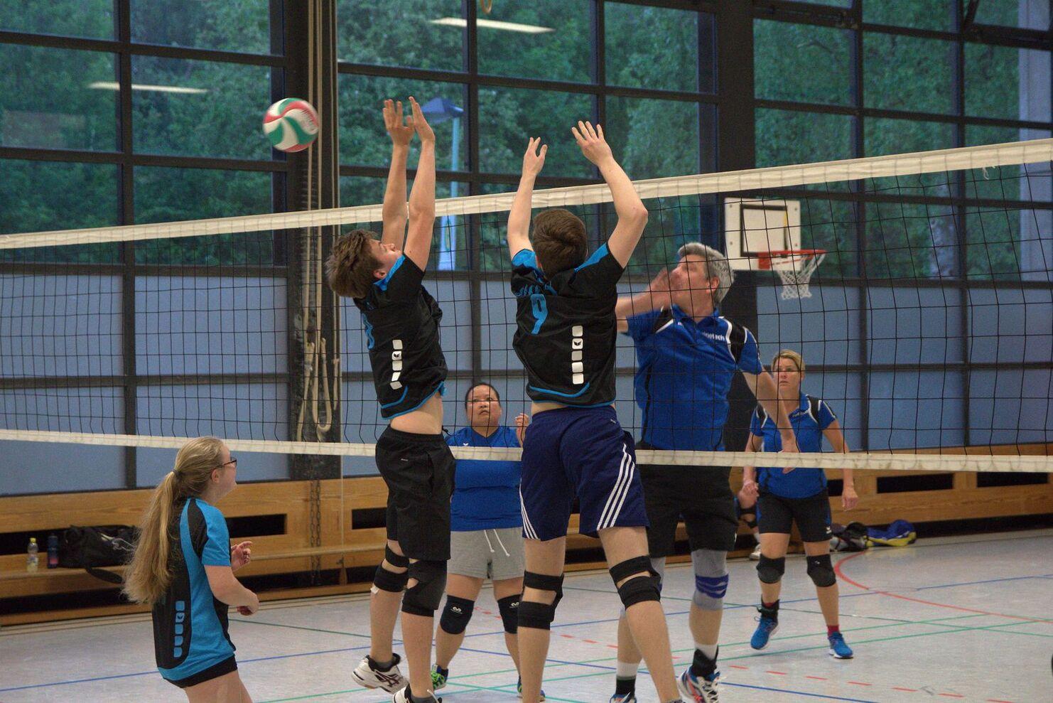 Zwei Spieler springen, um einen Volleyball über das Netz zu blocken, während weitere Spieler im Hintergrund agieren.