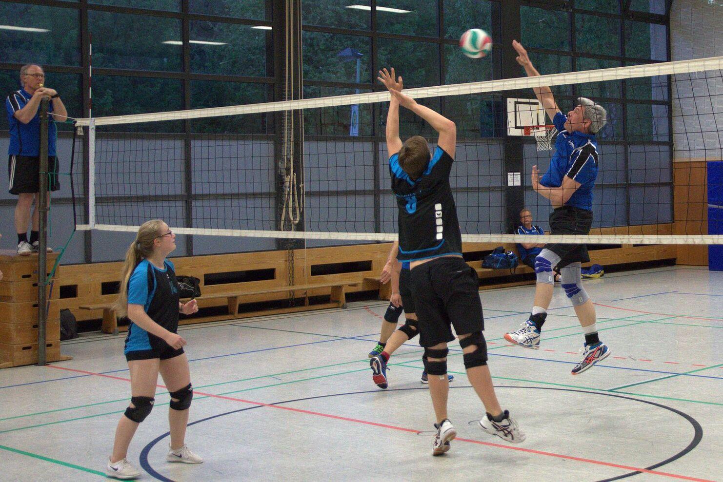 Volleyballspiel in der Halle: Spieler springen, um einen Ball über das Netz zu schlagen; Schiedsrichter beobachtet vom Turm aus.