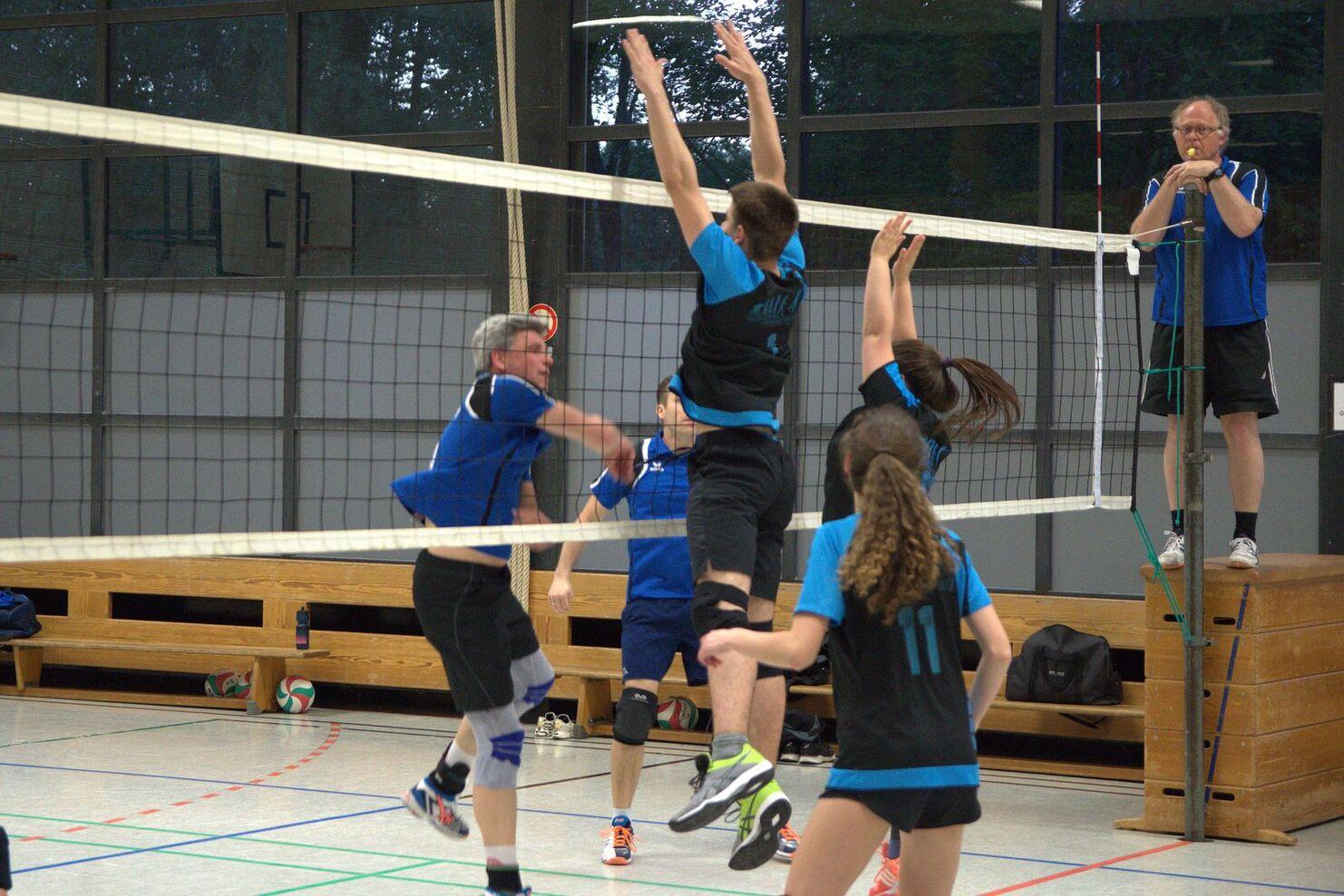 Zwei Volleyballspieler springen im Wettkampf über das Netz, während ein Schiedsrichter die Aktion beobachtet.