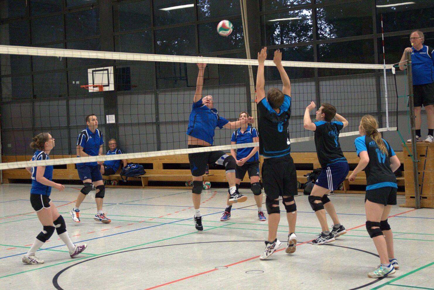 Volleyballspiel in der Halle: Spieler springen, um einen Ball über das Netz zu blocken, während andere zuschauen.