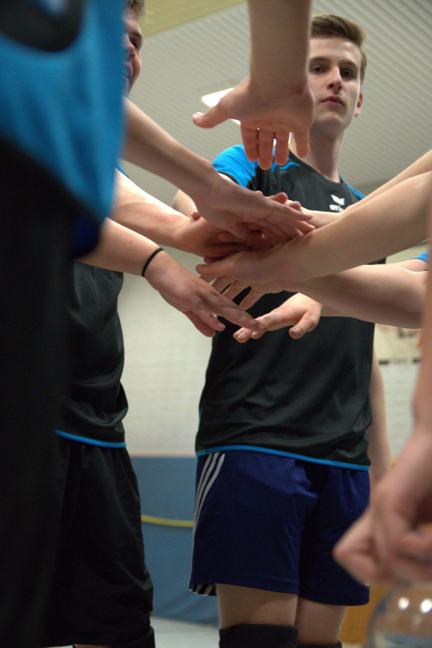 Hände von Spielern in einem Teamkreis, eine Person schaut konzentriert in die Kamera. Sportkleidung in Schwarz und Blau.