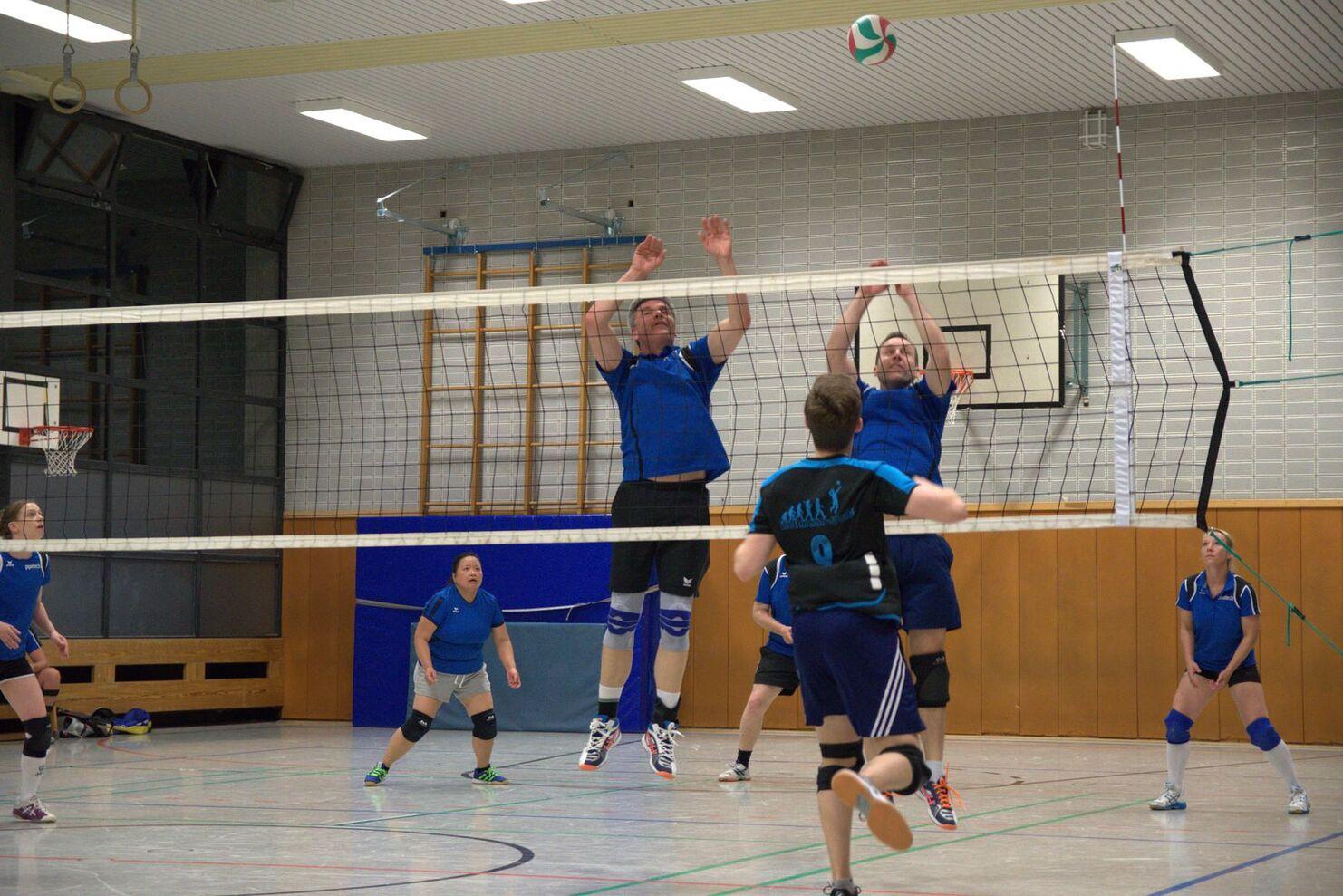 Volleyballspiel in einer Halle mit Spielern in blauen Trikots, die einen Ball über das Netz anvisieren.