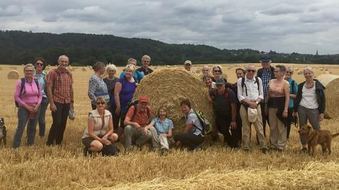 Gruppe von Wanderern posiert vor Heuballen auf einem Feld unter bewölktem Himmel. Zwei Hunde sind ebenfalls sichtbar.