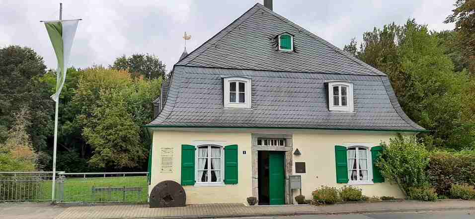 Goldberger Mühle in Mettmann Man sieht das Gebäude der Goldberger Mühle. Die Farben des Hauses sind hellgelb und grau. Links davon steht ein Fahnenmast.