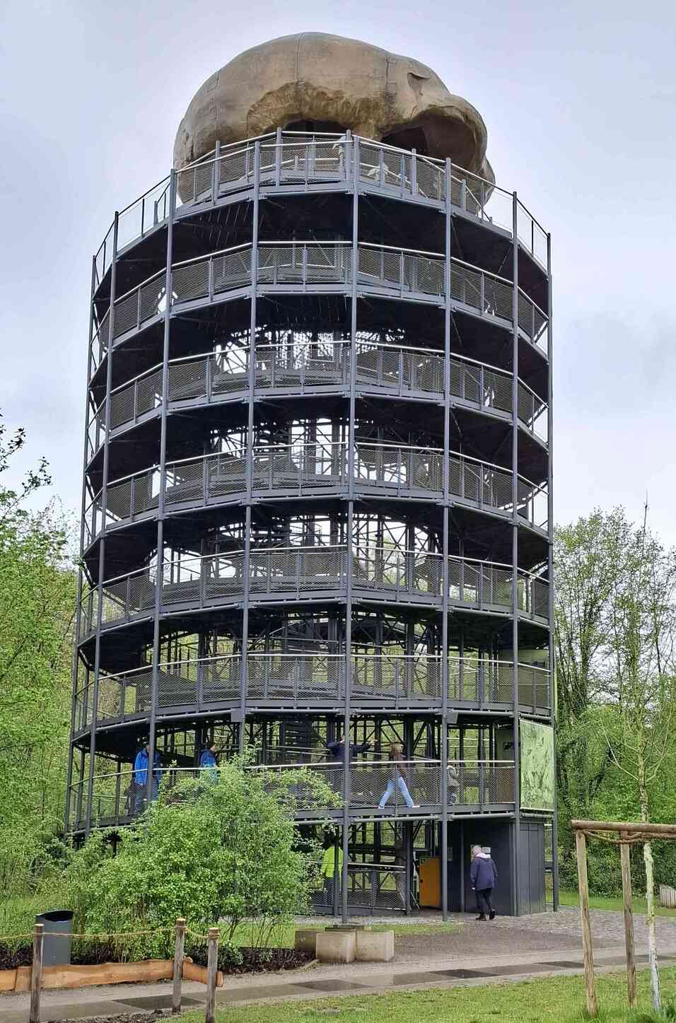 Aussichtsturm mit einer großen Skulptur eines Schädels auf der Spitze, umgeben von Bäumen und Besuchern auf den Plattformen.