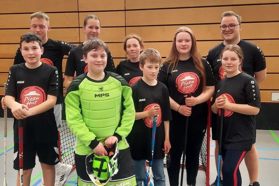 Gruppenbild von neun Jugendlichen in sportlicher Kleidung mit Schlägern und Torwartausrüstung in einer Sporthalle.
