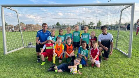 Gruppe von Kindern in Fußballtrikots posiert lächelnd vor einem Tor auf einem Rasenplatz, trainiert von zwei Erwachsenen.
