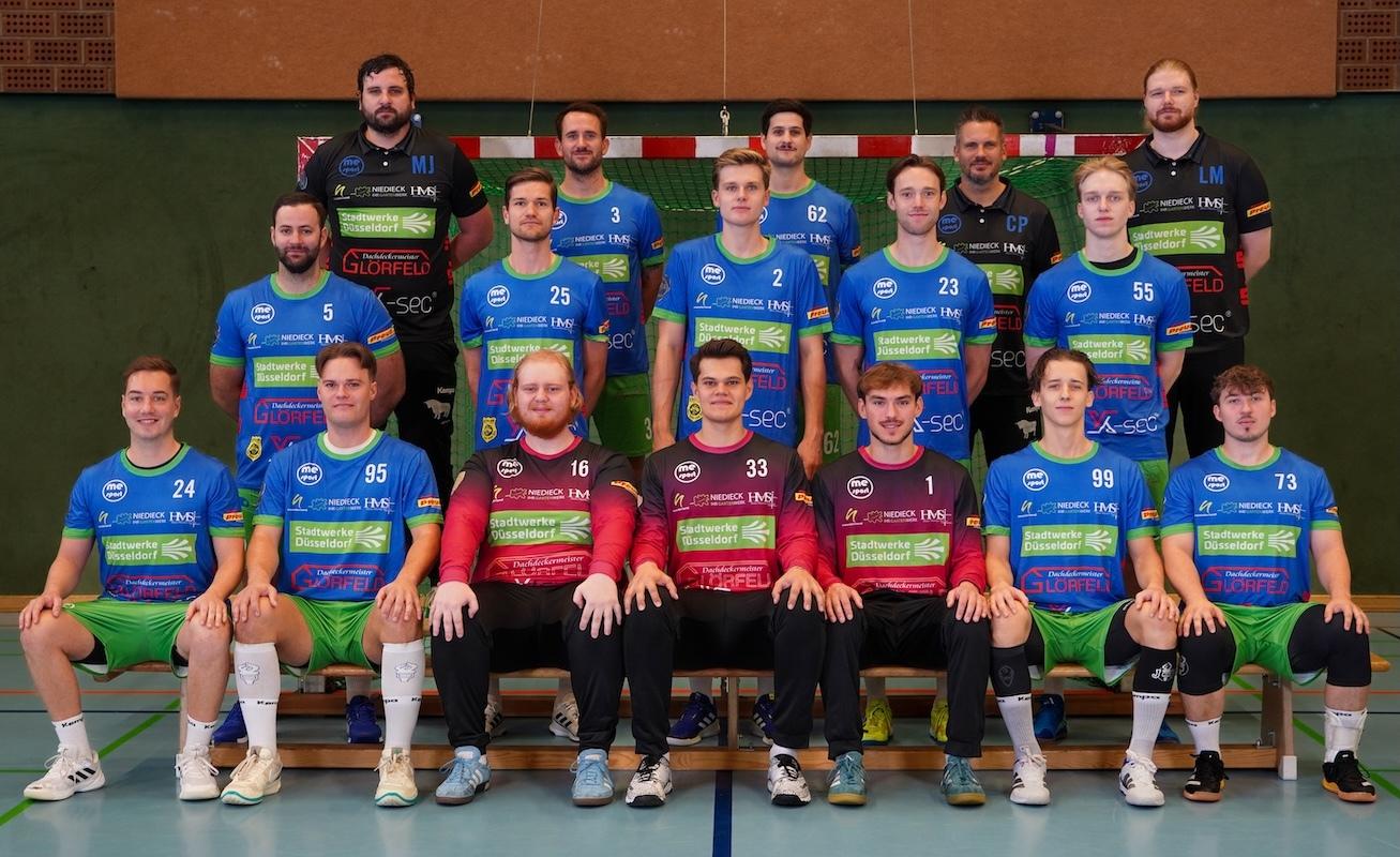 Mannschaftsfoto einer Handballmannschaft in blauen und roten Trikots auf einer Sporthalle-Bank mit Handballtor im Hintergrund.