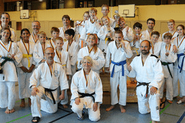 Gruppe von Judokas in Weiß und Farben, posierend nach einem Wettkampf mit Medaillen und Trophäen in einer Sporthalle.