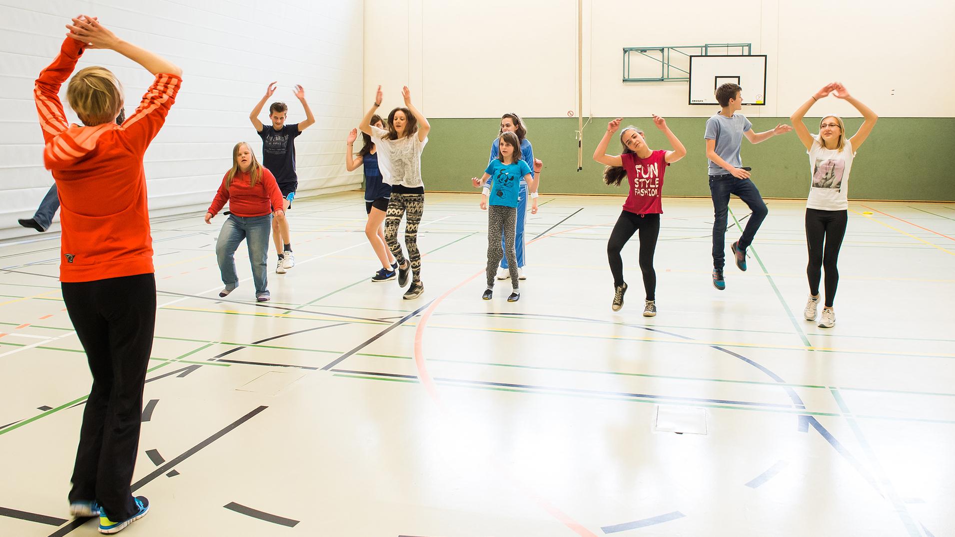 Gruppentraining in einer Sporthalle: Jugendliche springen und tanzen unter Anleitung einer Trainerin in Sportkleidung.