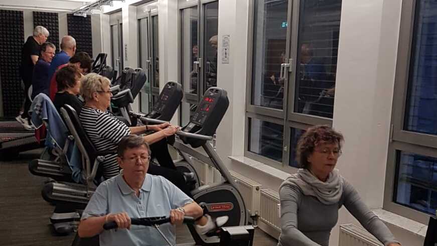 Gruppe älterer Personen beim Rudertraining an Geräten in einem modernen Fitnessraum mit großen Fenstern.