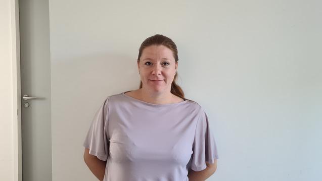 Frau mit langen Haaren steht lächelnd an einer Wand in einem hellen Raum, neben einer geschlossenen Tür und einem Feuerlöscher.