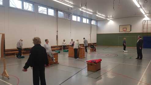 Gruppen von Senioren trainieren in einer Sporthalle, umgeben von Turngeräten und Matten.