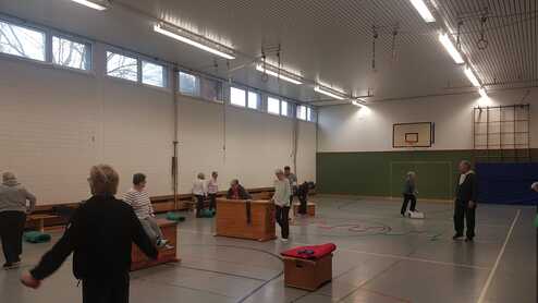 Ältere Erwachsene trainieren in einer Sporthalle mit Kästen, Matten und Bändern in einer aktiven Gruppenübung.