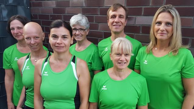 Gruppe von sieben Personen in grünen Sporttrikots, stehend vor einer Mauer, freundlich lächelnd.