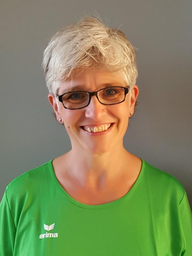 Frau mit kurzem, grauem Haar, Brille und grünem Shirt lächelt vor einem grauen Hintergrund.