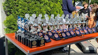 Tisch mit Pokalen und Medaillen, umgeben von Besuchern bei einer Veranstaltung im Freien.