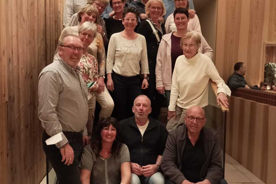 Gruppenselfie von 24 Personen in einem modernen Innenraum auf einer Treppe mit Holzverkleidung.