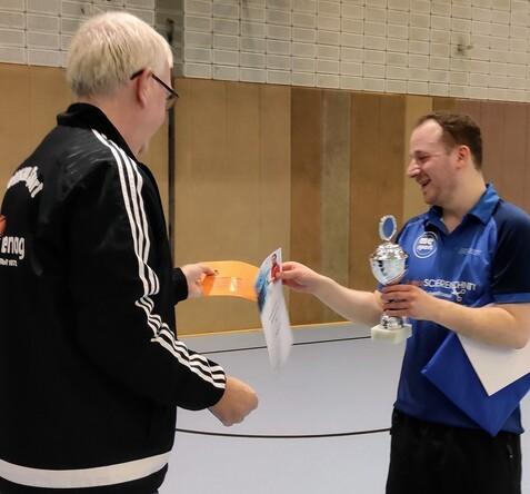 Zwei Personen in Sportkleidung, eine überreicht einen Pokal und ein Zertifikat an die andere in einer Sporthalle.