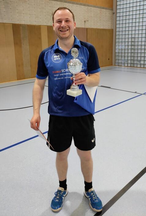 Lächelnder Spieler hält einen Pokal und eine Urkunde in einem Sporthalle mit blauen Linien auf dem Boden.