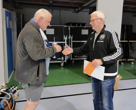 Zwei Männer in einem Sportraum schütteln sich die Hände; einer überreicht einen Pokal.