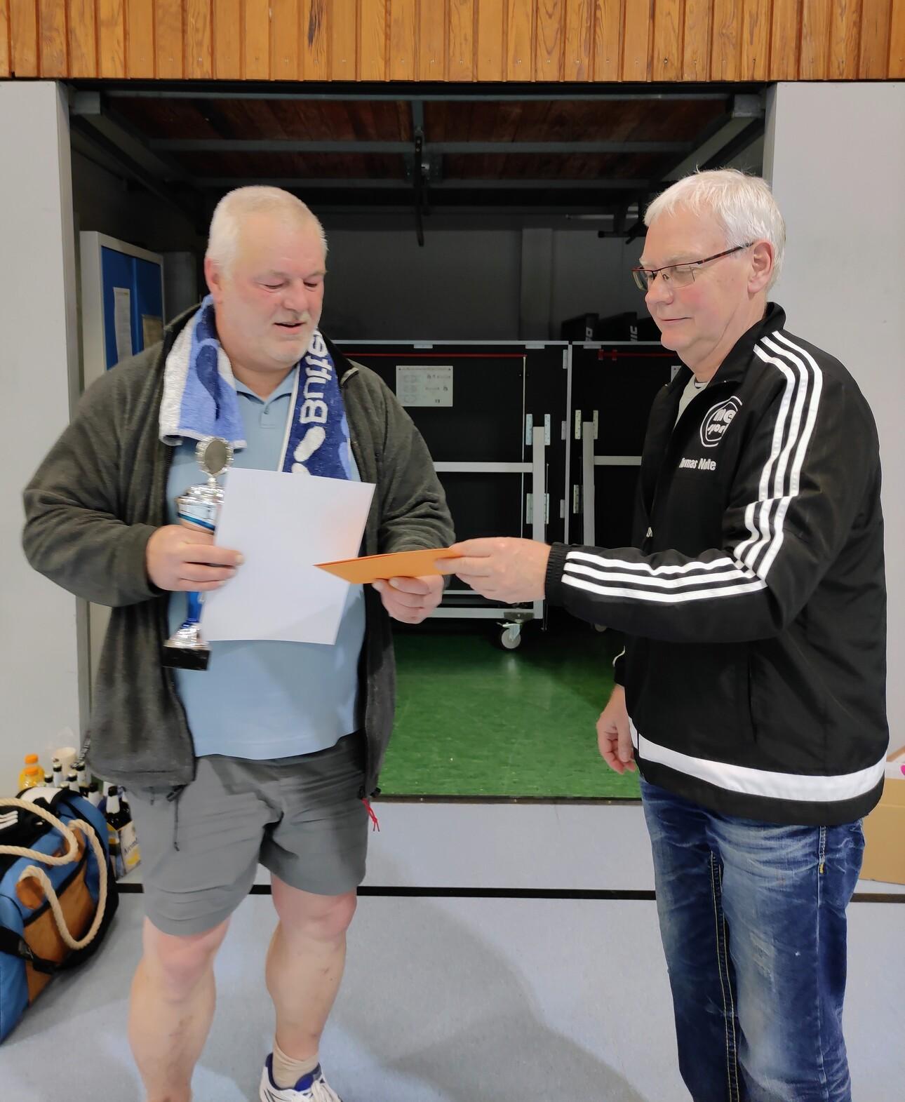 Zwei Männer in einer Sporthalle überreichen sich eine Urkunde und einen Pokal. Der Mann links trägt ein Handtuch um den Hals.