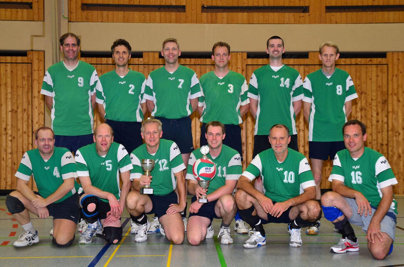 Mannschaftsfoto einer Volleyballgruppe in grünen Trikots, mit Pokalen, in einer Sporthalle.