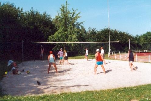 TSV Beachanlage Gruppe von Personen spielt Beachvolleyball auf einem Sandplatz im Freien, umgeben von Bäumen und Sportanlagen.