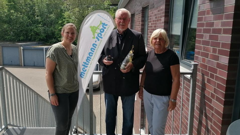 Drei Personen stehen draußen vor einem Gebäude, halten eine Auszeichnung und posieren neben einem sportlichen Banner.