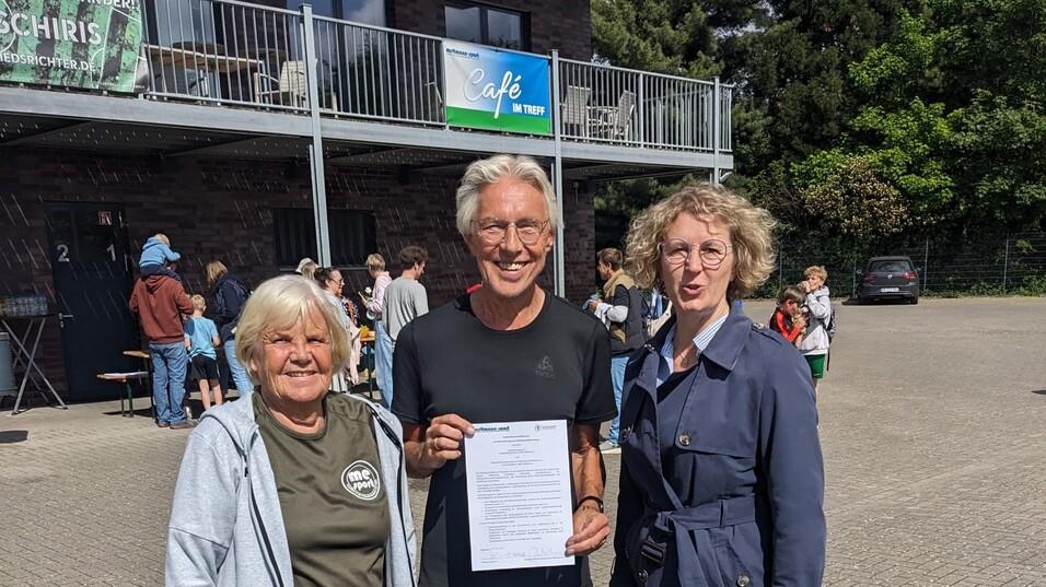 Drei Personen stehen vor einem Gebäude, einer hält ein Dokument, im Hintergrund sind Menschen und Bänke zu sehen.