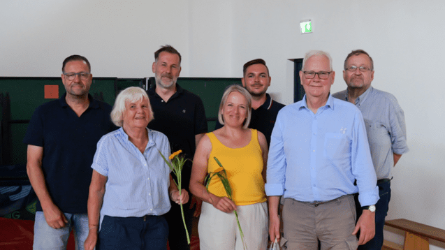 Gruppe von sieben Personen in einer Sporthalle, einige halten Blumen und Geschenke, beide Geschlechter vertreten.
