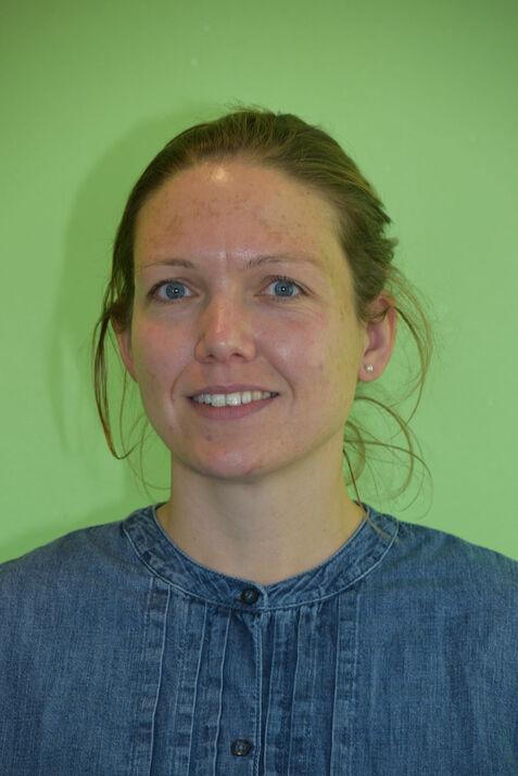 Porträt einer Frau mit blauen Augen und kurzen, lockigen Haaren, die ein Jeanshemd trägt, vor einer grünen Wand.