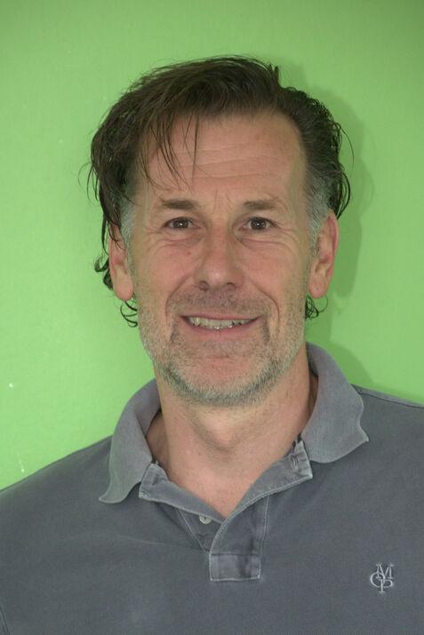 Nahaufnahme eines Mannes mit einem grauen Polo-Shirt vor einer grünen Wand, lächelnd und mit leicht zerzausten Haaren.