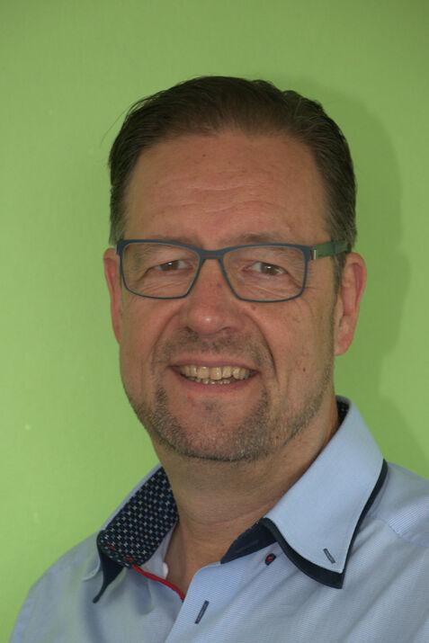 Mann mit Brille und Bart lächelt vor grünem Hintergrund, trägt ein hellblaues Hemd mit einem gemusterten Kragen.