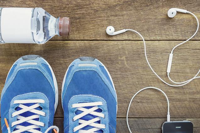 Blick von oben auf blaue Sportschuhe, Wasserflasche, Kopfhörer und ein Smartphone auf einer Holzoberfläche.