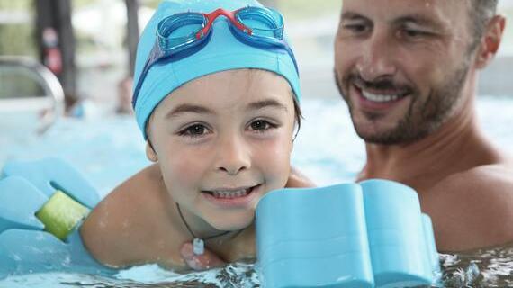 Ein Junge mit blauer Schwimmmütze und Schwimmhilfen lächelt im Wasser, während ein Mann hinter ihm schaut und lächelt.