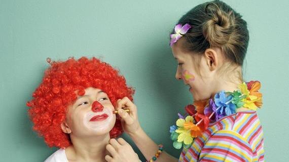 Kind mit rotem Lockenperücke und Gesichtsmalerei wird von einem Mädchen mit Blumenkranz geschminkt, vor einem grünen Hintergrund.