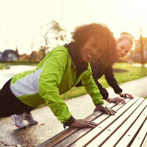 Zwei Frauen in Sportkleidung machen im Park Liegestütze auf einer Holzbank, die Sonne scheint.