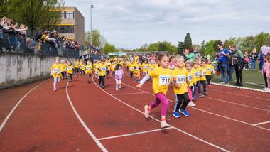 Eine Gruppe von Kindern in gelben T-Shirts läuft auf einer Laufbahn während eines Sportevents, Zuschauer stehen ringsum.