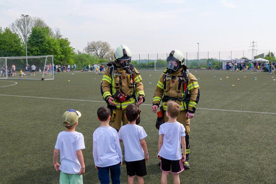 Zwei Feuerwehrleute in Schutzanzügen sprechen mit mehreren Kindern auf einem Sportplatz. Im Hintergrund sind Zuschauer und Aktivitäten sichtbar.