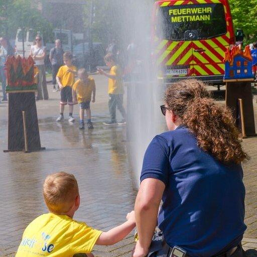 Kind im gelben T-Shirt sitzt auf dem Boden und blickt mit einer Feuerwehrfrau auf einen Wasserstrahl aus einem Feuerwehrschlauch.