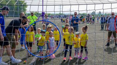 Minisportabzeichen Gruppenspiel von Kindern in gelben T-Shirts auf einem Sportplatz mit einer blauen Zielscheibe im Vordergrund.
