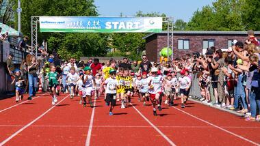 Start eines Kinderlaufes mit vielen laufenden Kindern auf einer roten Laufbahn, umgeben von jubelnden Zuschauern.